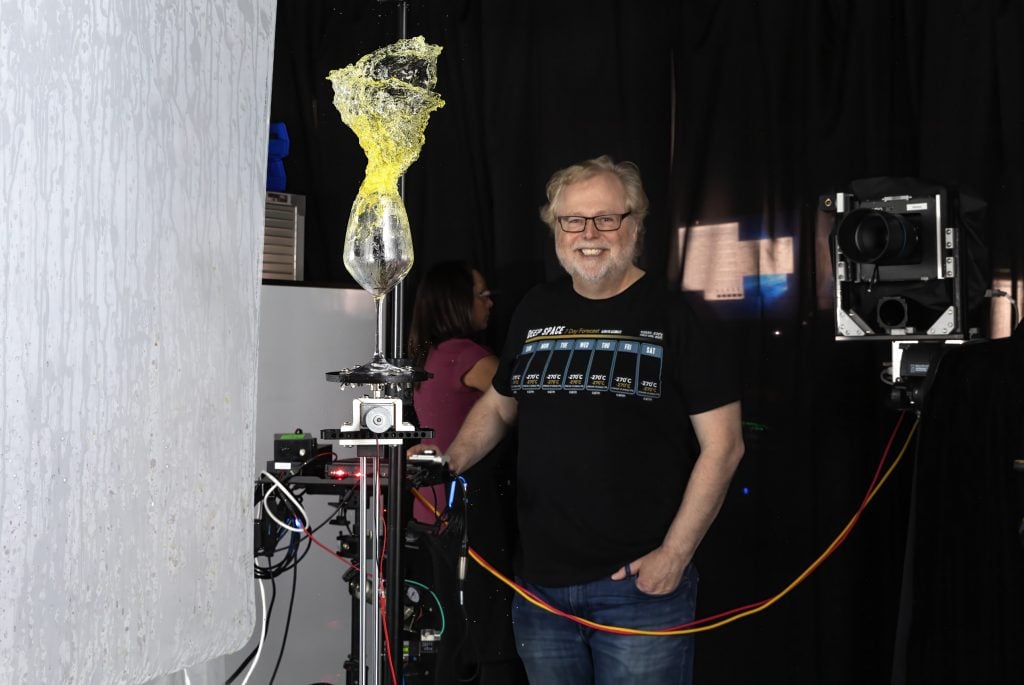 Nathan Myhrvold taking one of his frozen-motion photographs of wine. A middle-aged man with glasses and light wavy hair and a graying beard smiles for the camera as another large box camera takes a photo of a glass of white wine where the liquid is flying upward, captured in such crisp detail that it looks like colored glass.