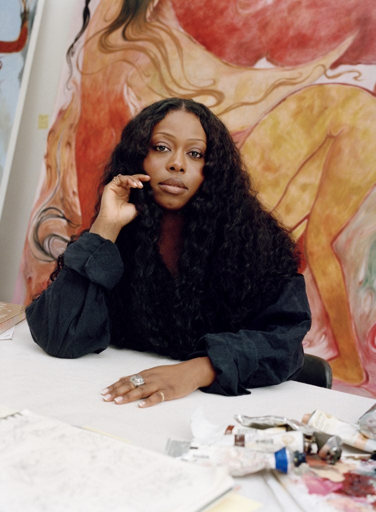 a black woman with long curly hair sits at a table in her art studio wearing a black button-down shirt