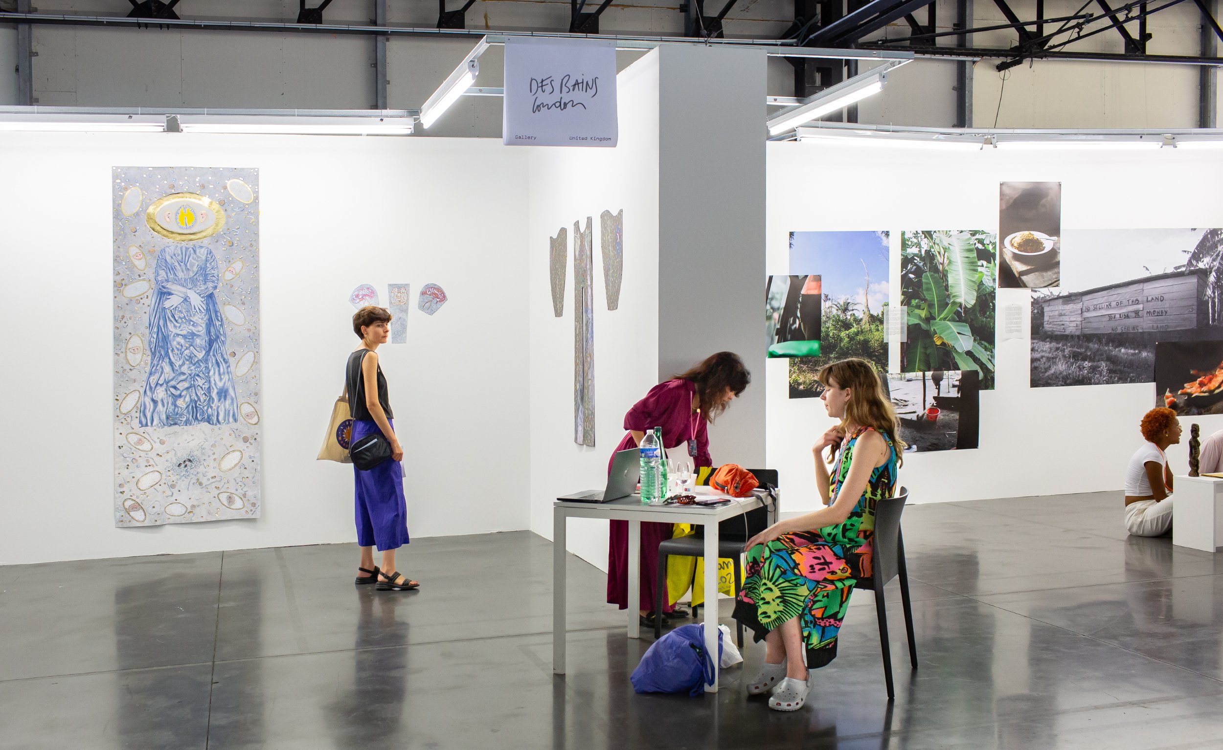 the interior of a fair with white walls partitioning two galleries with two well dressed people sitting at a table with a laptop