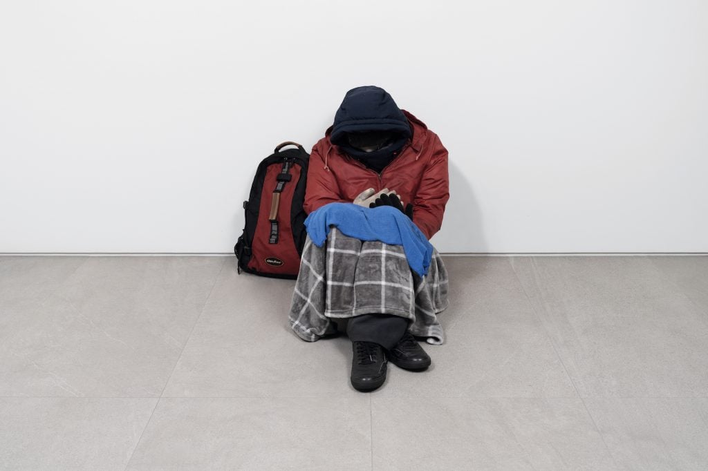 an image of a homeless figure huddled in a blanket with a knapsack nearby