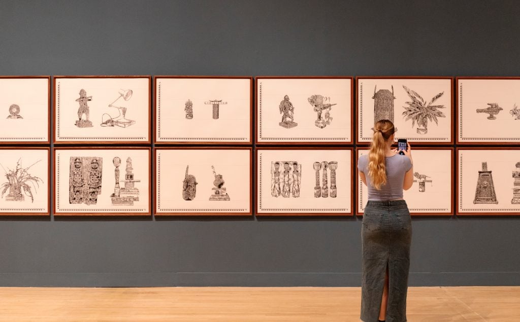 a photograph of a gallery space in which there are framed intricate drawings in black on white of ancient cultural objects side by side with everyday contemporary objects from the home 