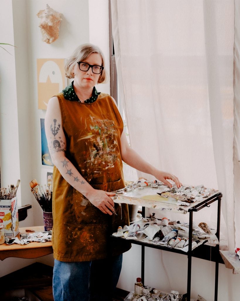 a photo of a woman in glasses and a smock in a studio with paints