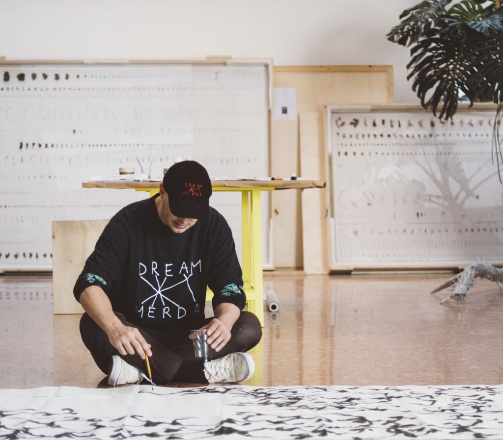 a man in a black outfit sits inside a spacious interior looking down with concentration so we can't see his face while he uses a brush to apply dark pigment in rhythmic patterns to a large white paper surface spread out on the floor 