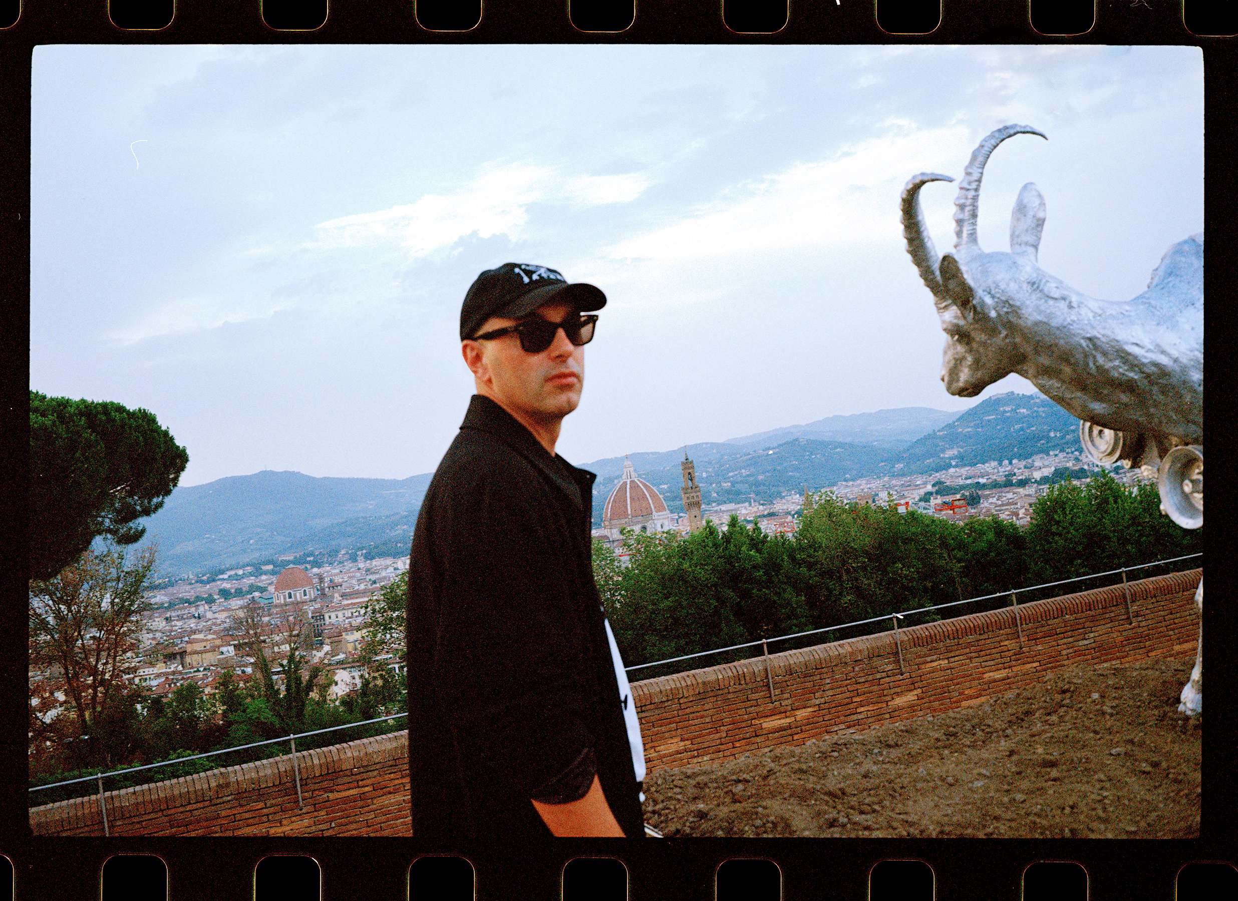 a man in a black shirt and a black cap and sunglasses stands in profile against a landscape, he turns to look at the photographer, to the right we can see the metallic silver head of a sculpture of a goat and in the background we can see the dome of an old Italian religious building