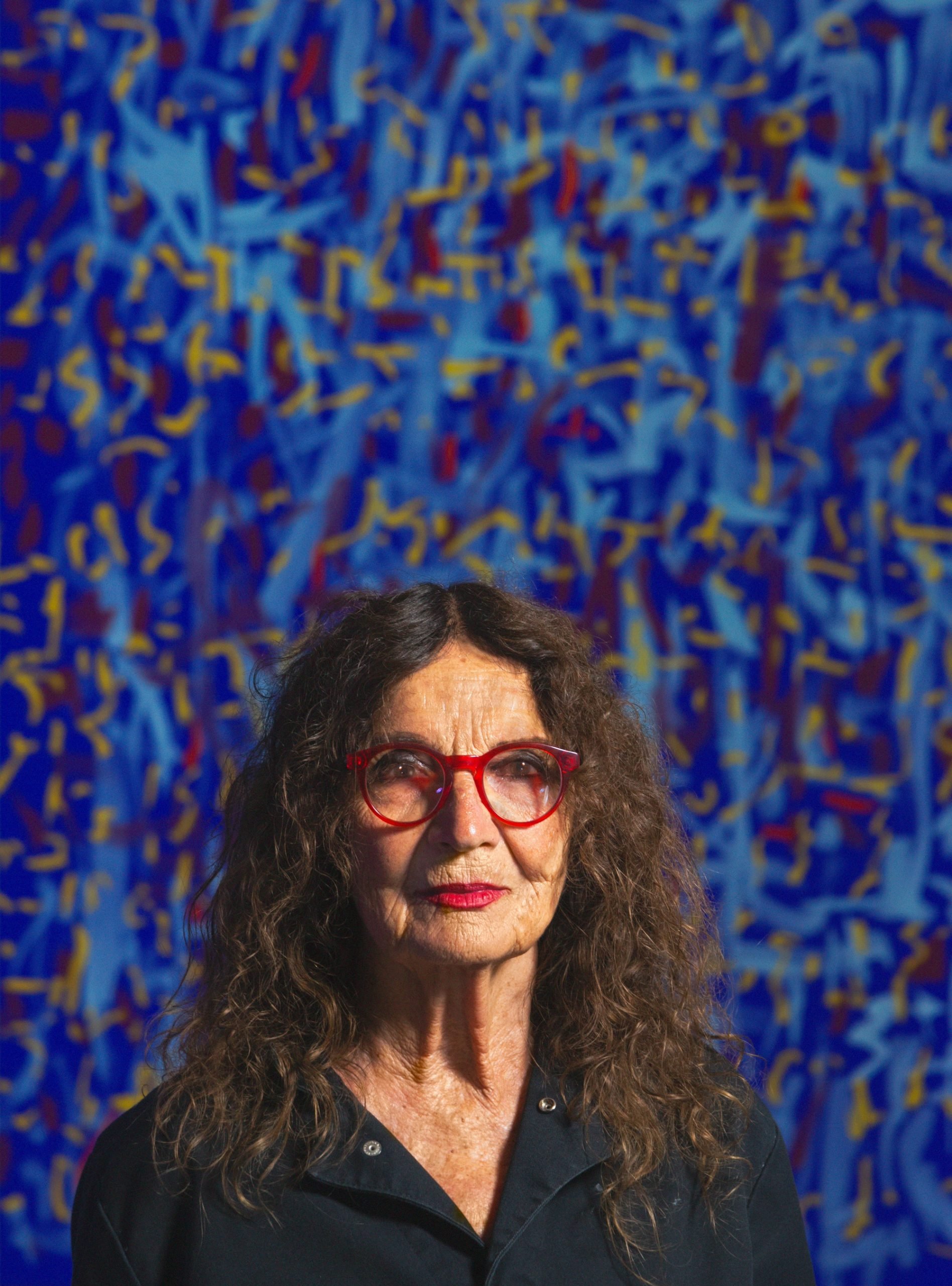 a woman in her 70s with brown curly hair and red glasses looks out at the camera. behind her is an abstract blue paintings