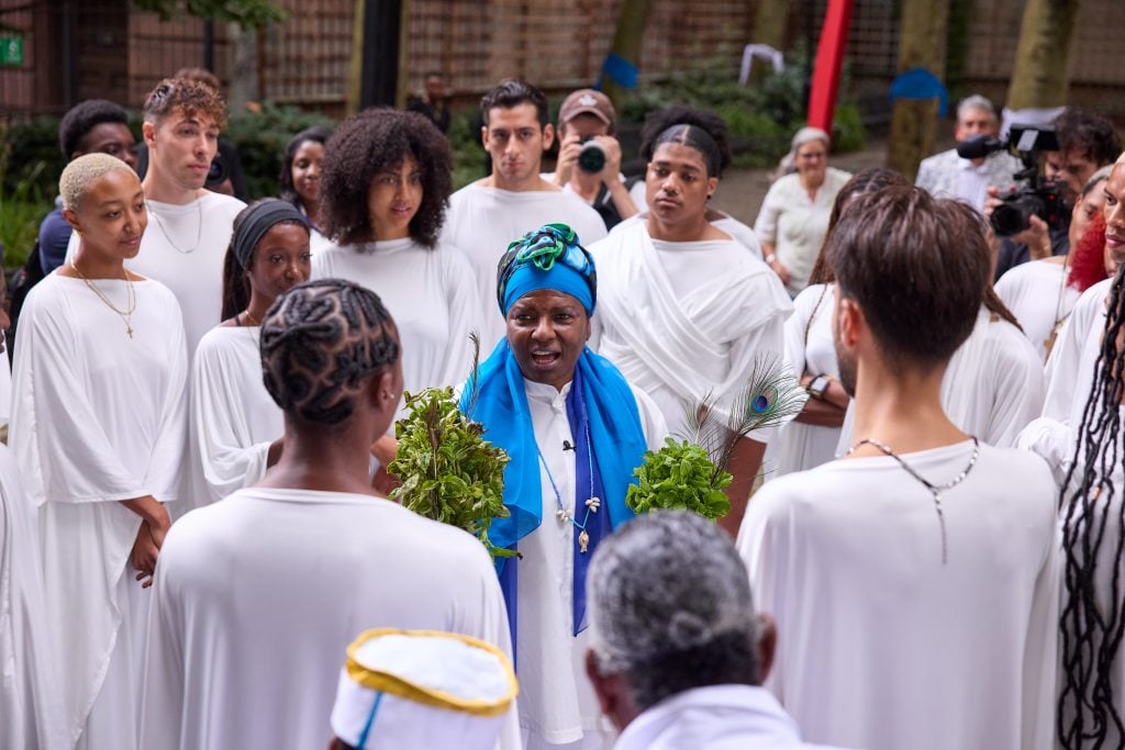 Artist María Magdalena Campos-Pons in a blue turban and white clothing speaks to participants in Harlem Art Park all dressed in white