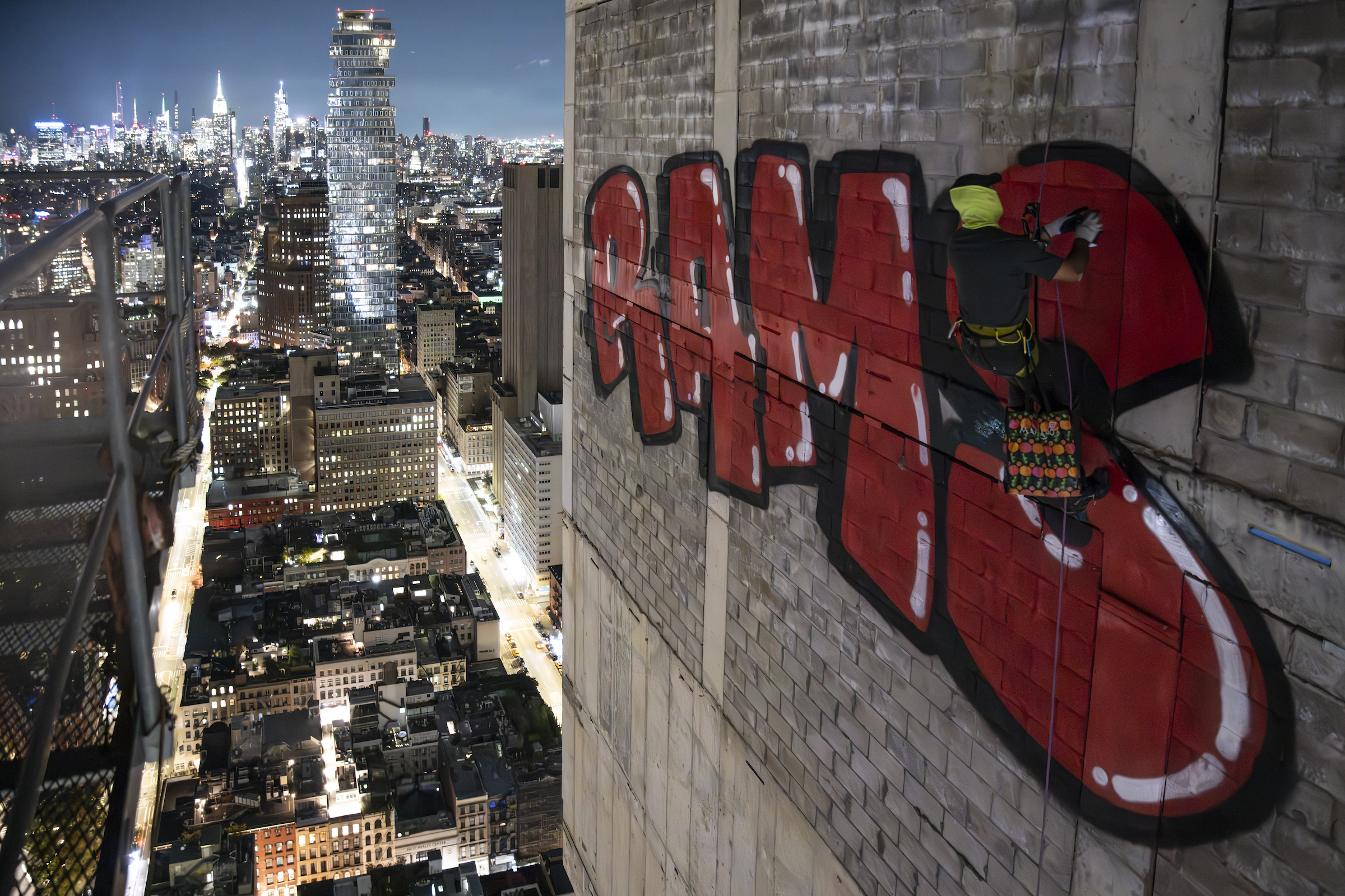 RAMS rappelling down the side of the abandoned skyscraper 45 Park Place to spray paint his name on the building in red. It is nighttime, and the New York City skyline can be seen behind him.