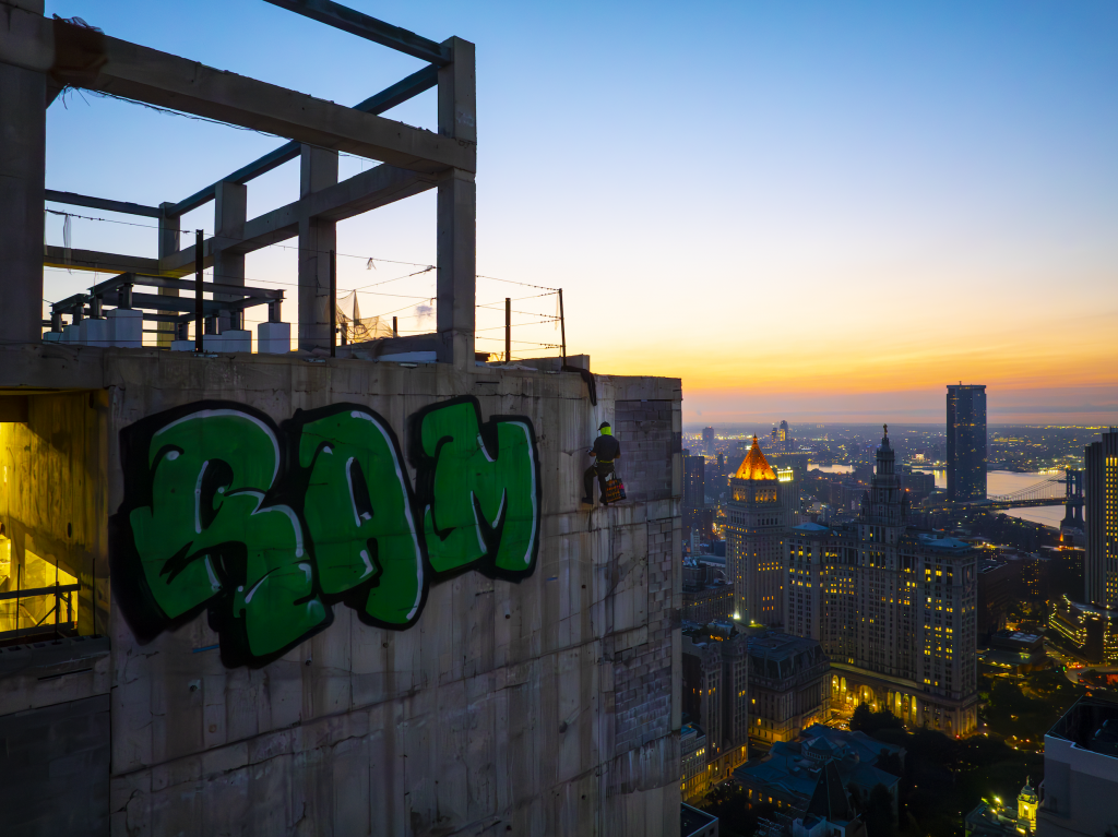 RAMS rappelling down the side of the abandoned skyscraper 45 Park Place to spray paint his name on the building in green. The sun is rising over the New York City skyline as he completes the piece.