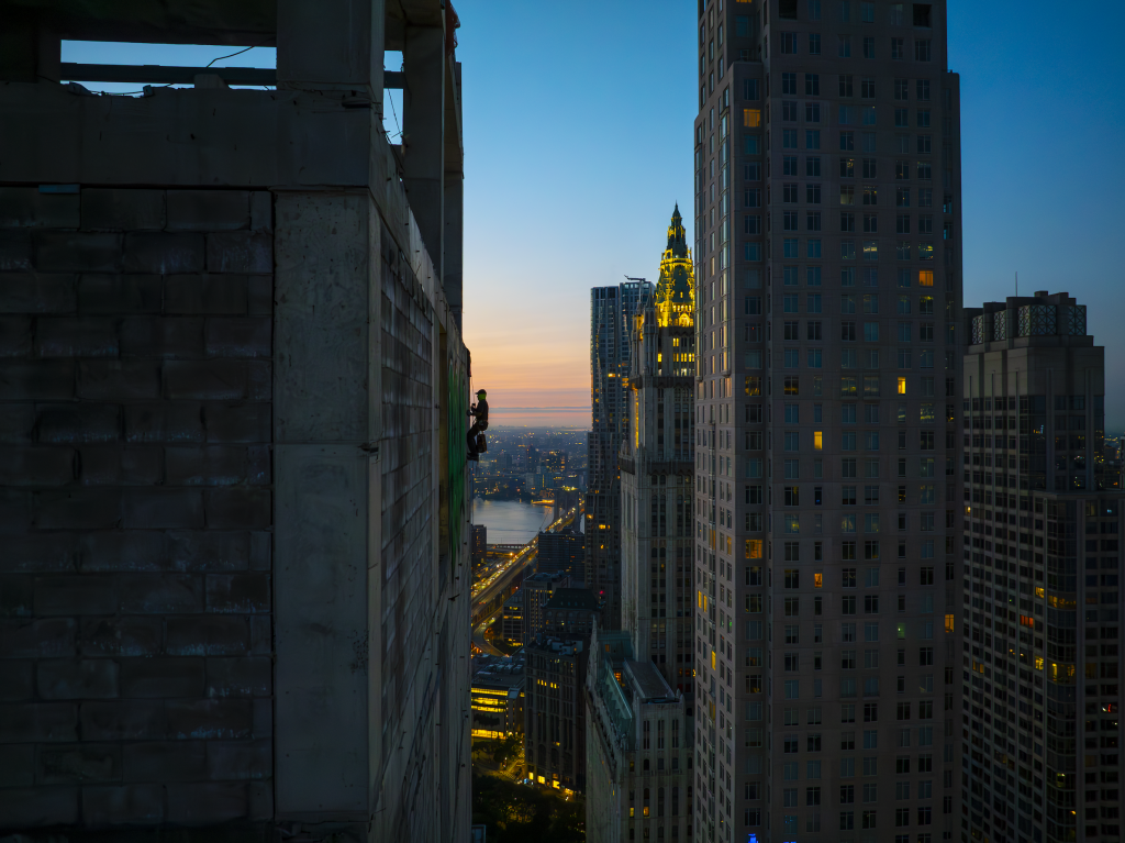 RAMS rappelling down the side of the abandoned skyscraper 45 Park Place to spray paint his name on the building. The early morning light can be seen, with the Woolworth Building and other skyscrapers behind. 