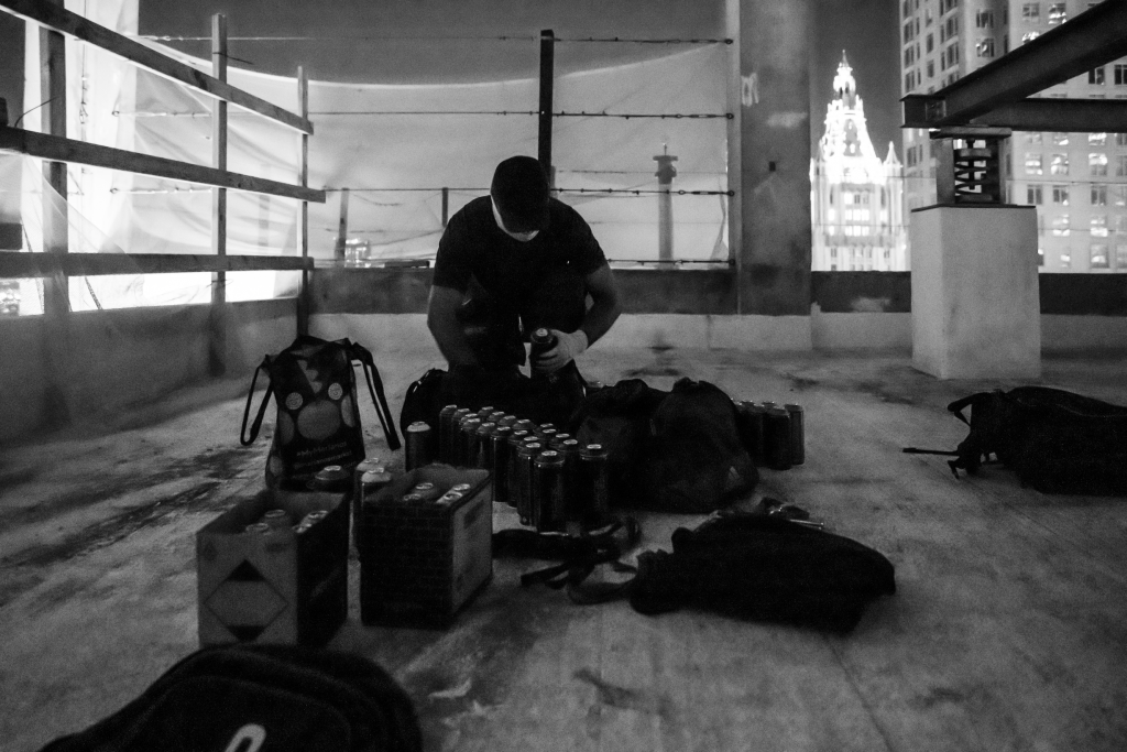 RAMS setting up to graffiti 45 Park Place. The shadowy black and white photo obscures the artist's face, showing him sitting on the floor of the construction site in the abandoned skyscraper getting out his paint and tools. A lit skyscraper can be seen behind him. 