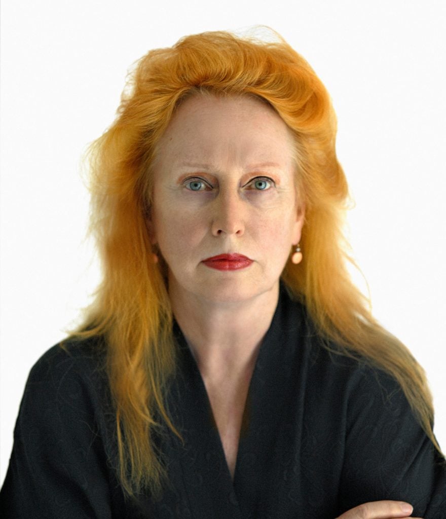 a woman with dyed ginger hair wearing a black shirt with red lipstick and earrings stares directly at the camera