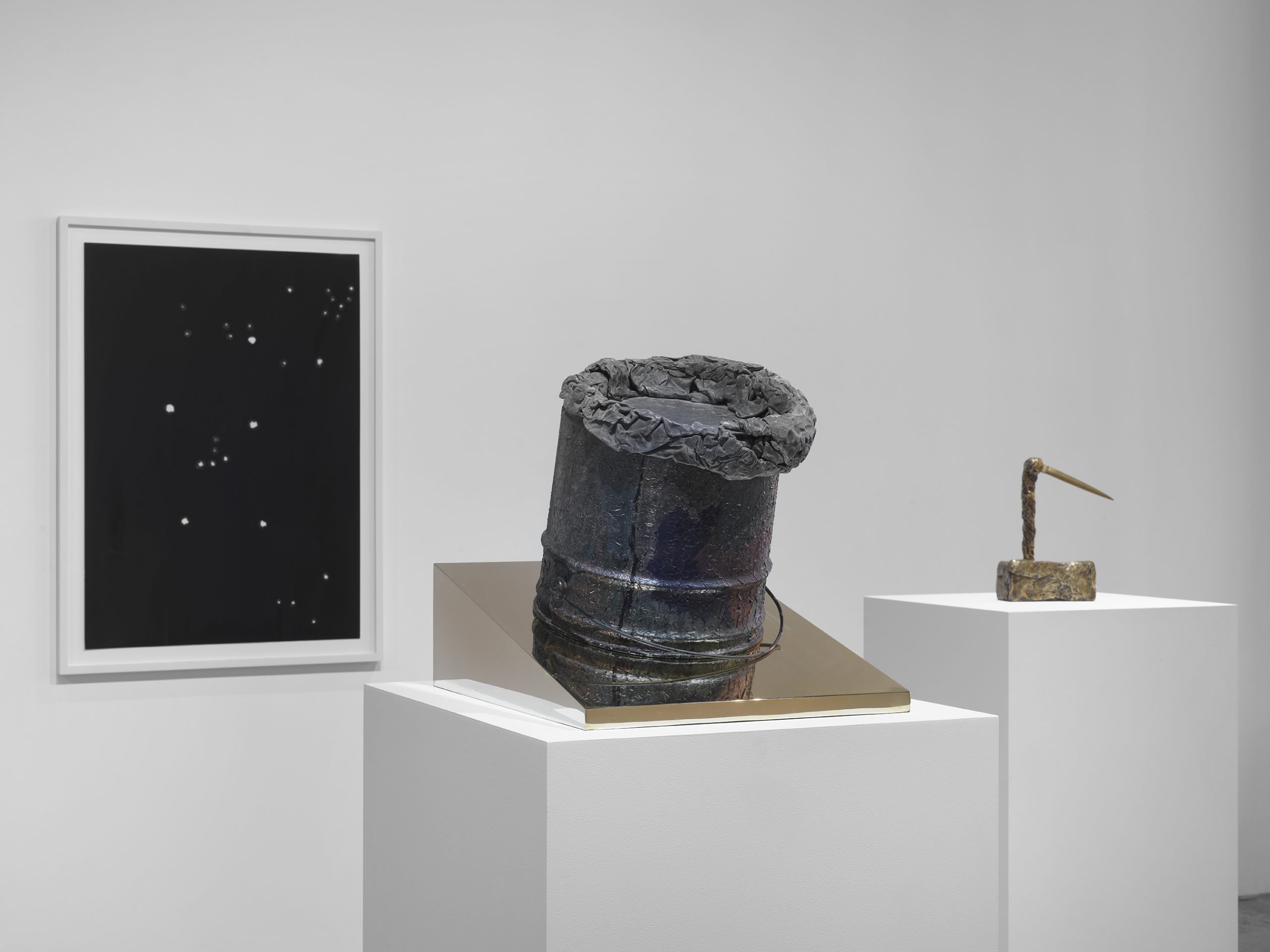 Two sculptures sit on white plinths and a black wall piece hangs on a white wall