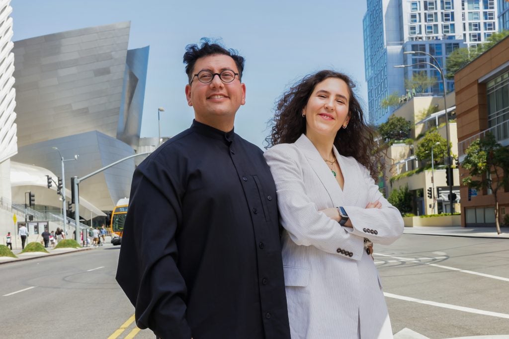 two people stand smiling in the middle of the road the sky is blue behind and there is s metal building to the left hand side