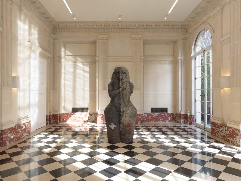 an imposing deity like sculpture has Egyptian and Native American components and stands in a very grand gallery space with a checkerboard floor 