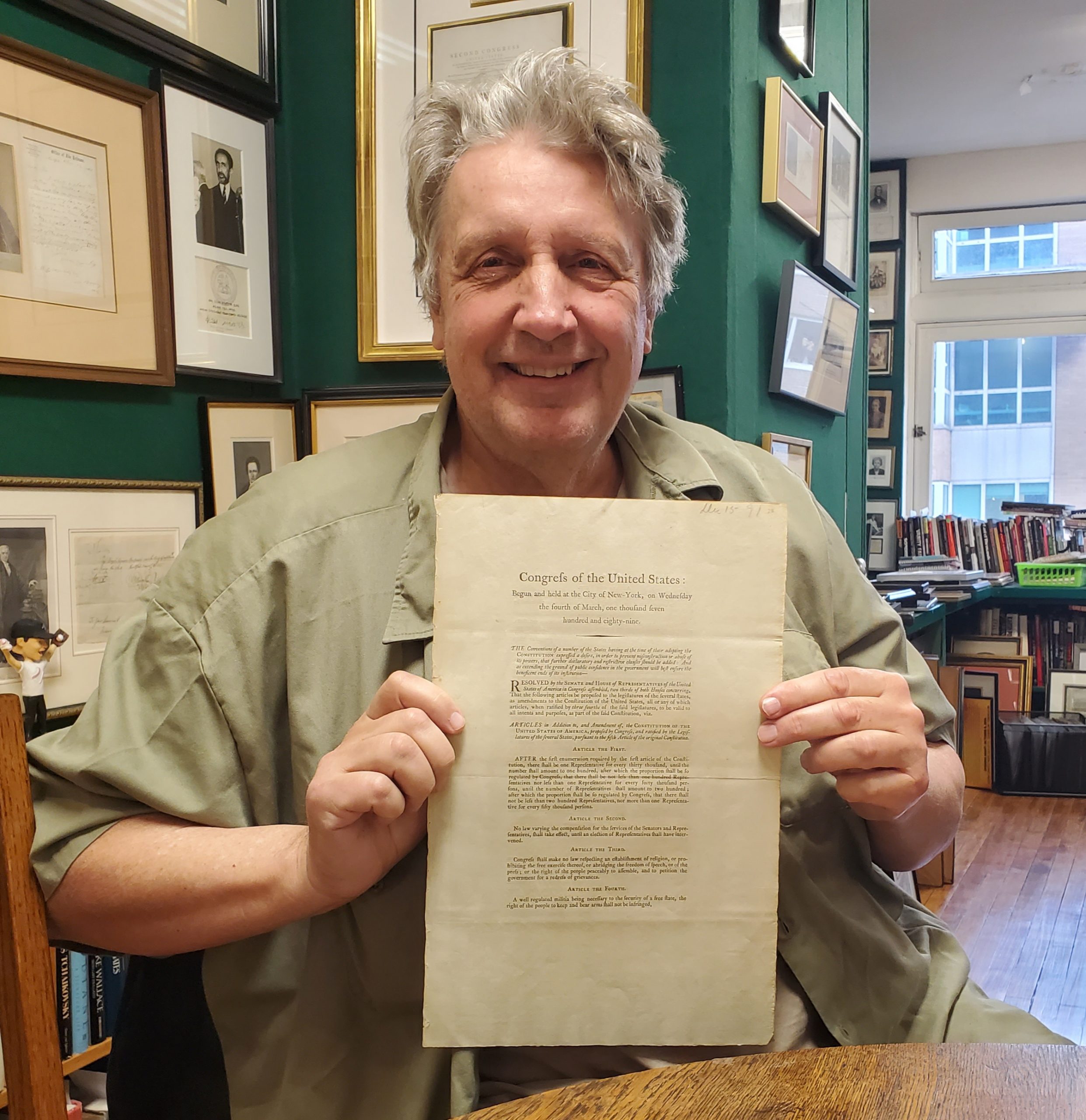 New Orleans historic document collector Stanley Klos with his copy of the 1789 Congressional Broadside printing of the 12 Articles, known as the Bill of Rights, passed by Congress on September 25, 1789. He is an older white man with gray hair, smiling and sitting in a room with numerous framed documents and photographs hanging on the walls of a home.