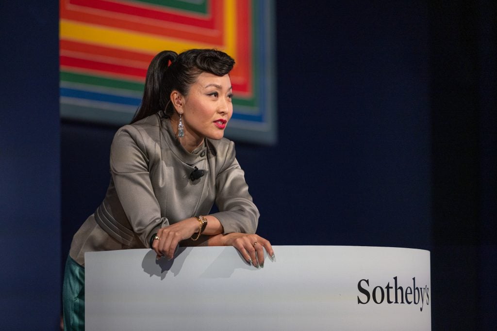 An asian woman in a ponytail leans over the rostrum in front of a rainbow colored geometric painting