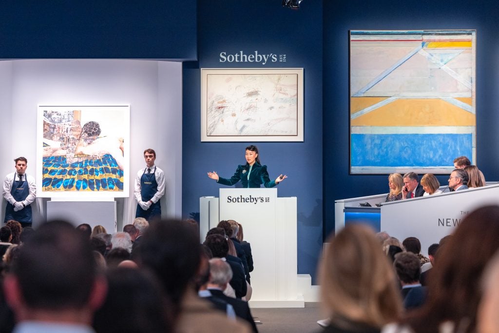 A wide-view of an art auction salesroom with an asian woman at the center with her arms outstretched taking bids.