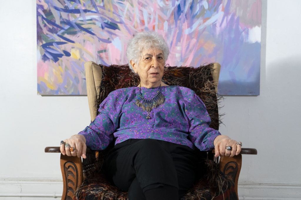 a woman in a purple top sits in an armchair in front of a large abstract painting in shades of purple, pink and blue