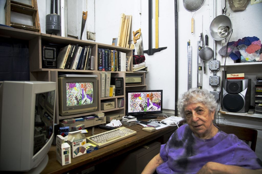 a photograph in which a woman is sitting in the foreground in a studio space but turns to look at the viewer, she is sitting at a desk that is filled with old fashioned box-shaped computer monitors as well as shelves of books and tools hanging along the walls