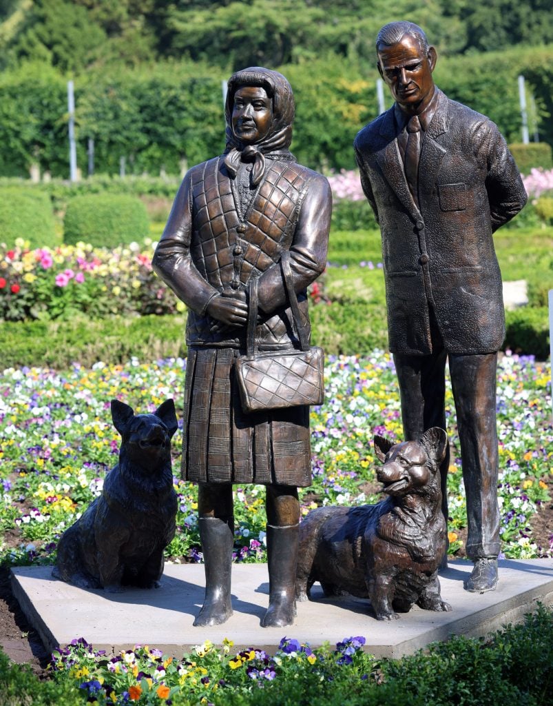 a green gardens setting against which stands two bronze figures, the woman is wearing a vest and a headscarf and the man is wearing a suit, at their feet are two Corgi dogs