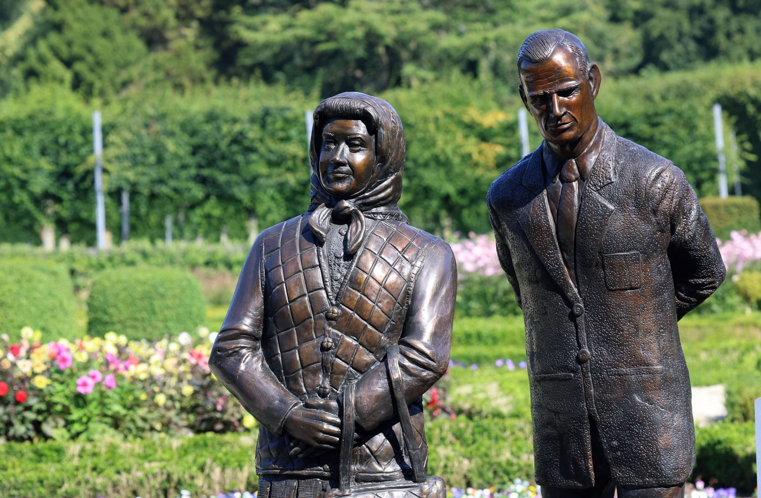 a green gardens setting against which stands two bronze figures, the woman is wearing a vest and a headscarf and the man is wearing a suit