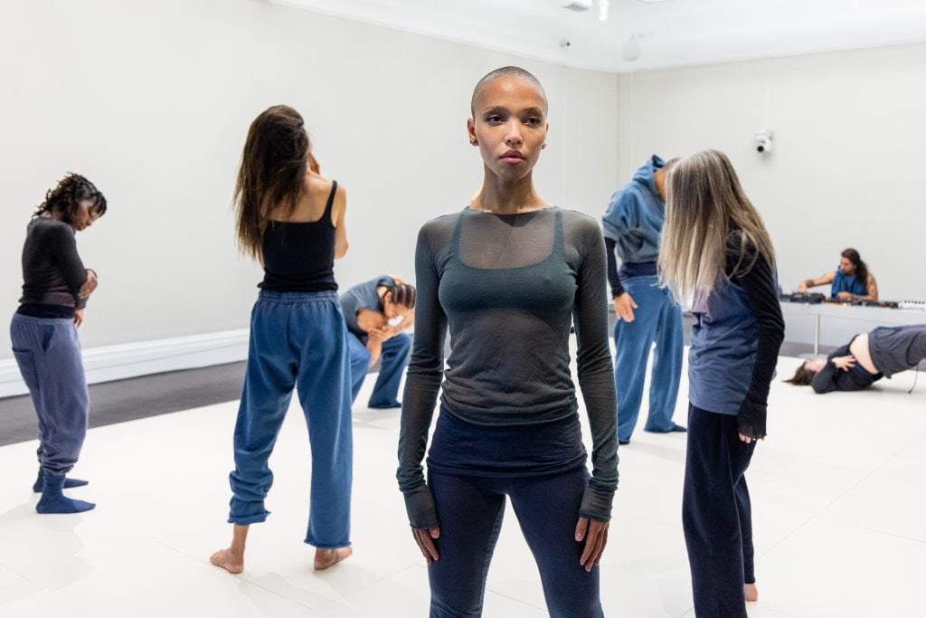 a woman stands in front of a dozen people in a white room