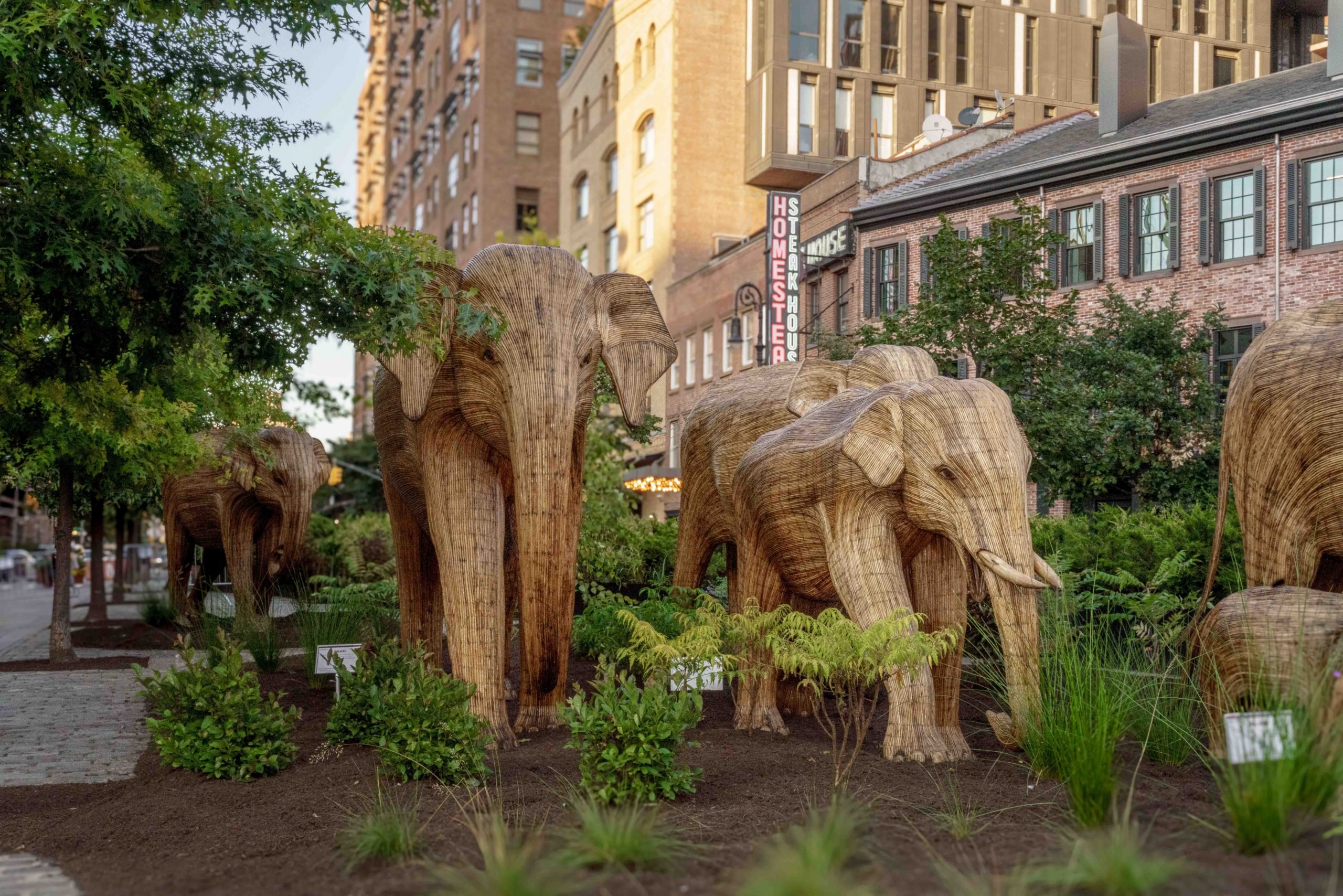 "The Great Elephant Migration" in New York's Meatpacking District. A herd of life-size wooden elephant sculptures amid greenery in front of brick buildings in the city.