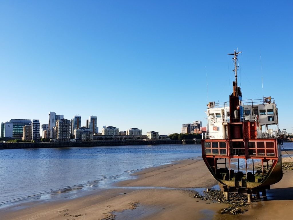 a large ship like structure that is clearly an artwork sits on the bank of a large strech of water, the River Thames, with high rise buildings of a city in the distance and a bright blue sky