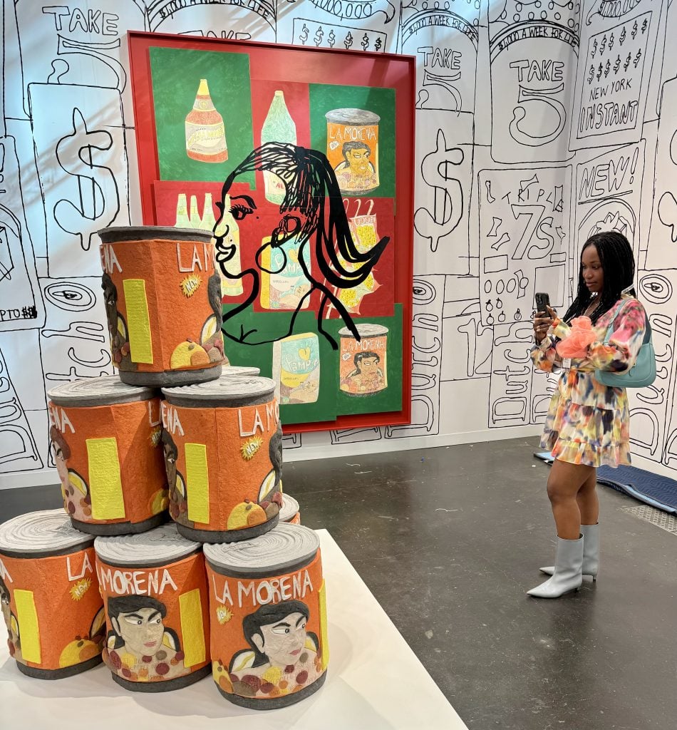 Tschabalala Self photographing her installation "Bodega Run" with Two Palms at the Armory Show. She is a young Black woman wearing a short, colorful print dress. There is a giant stack of cast paper La Morena can sculpture, and a collaged silkscreen work with a woman's profile over illustrations of bodega products. The wallpaper is black and white line drawings of more bodega goods. 