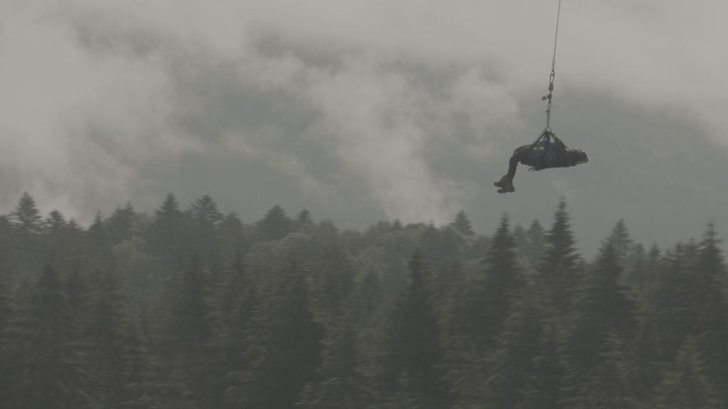 a dark grey sene in which an inert, human figure lying down can be seen suspended from a rope and floating over the tops of forest trees against a foreboding cloudy sky