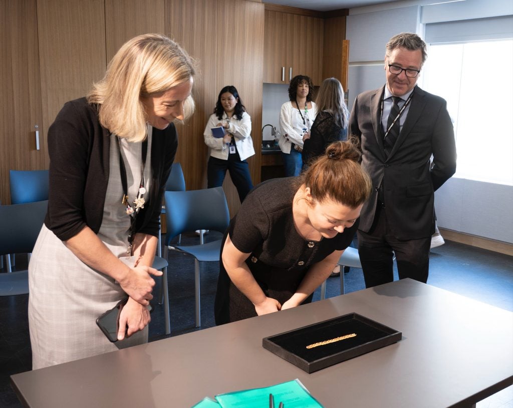 three people stand around a small necklace in a black case, they smile and one woman in the middle bends down to look closer, they are in a professional interior with some people lingering in the background