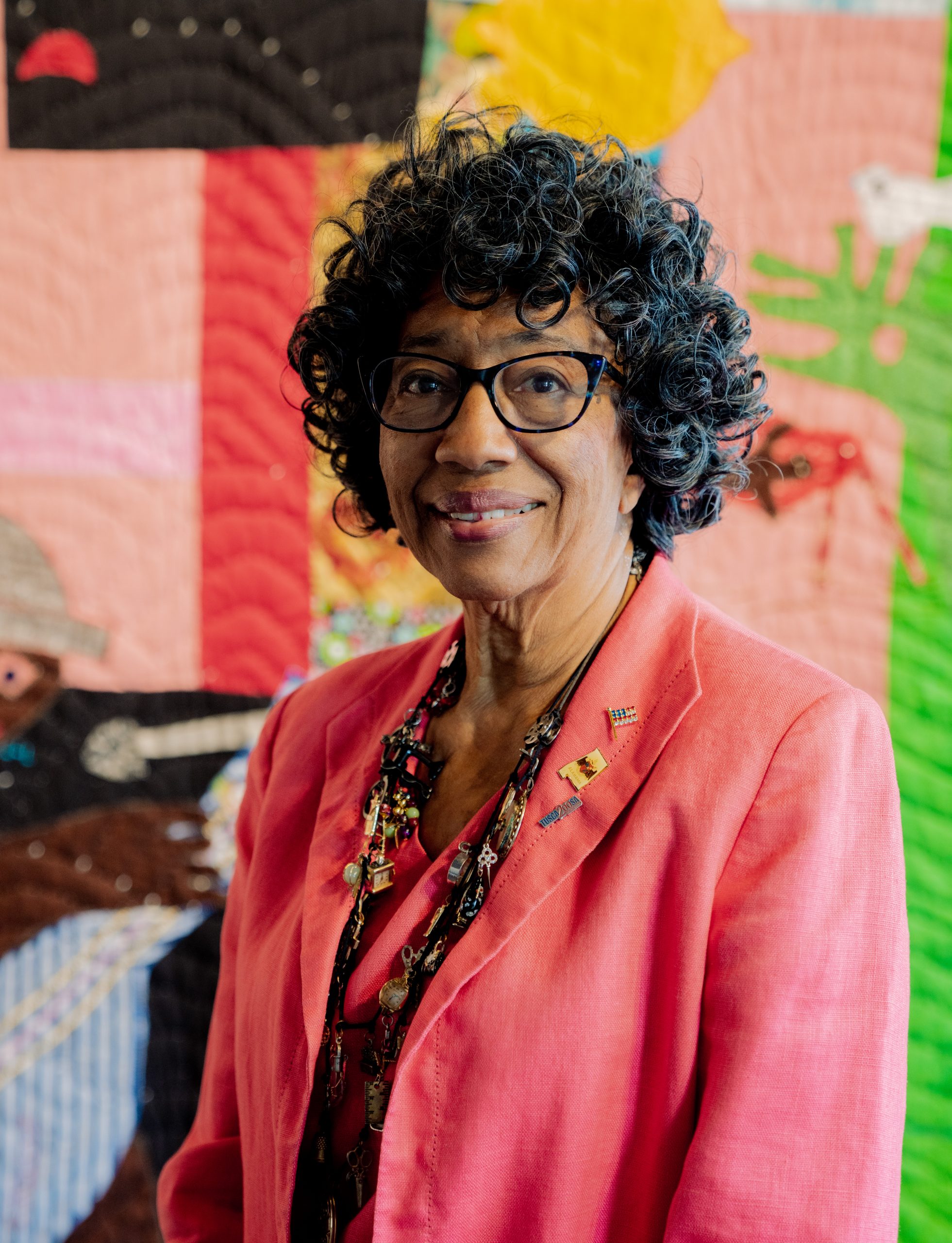 a photograph of an older black woman wearing gladdes and a coral pink suit. She is seated in front of a quilt