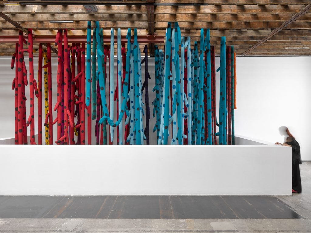 A view standing at a balcony looking at a large installation work made of stuffed fabric tentacles suspended from the ceiling.