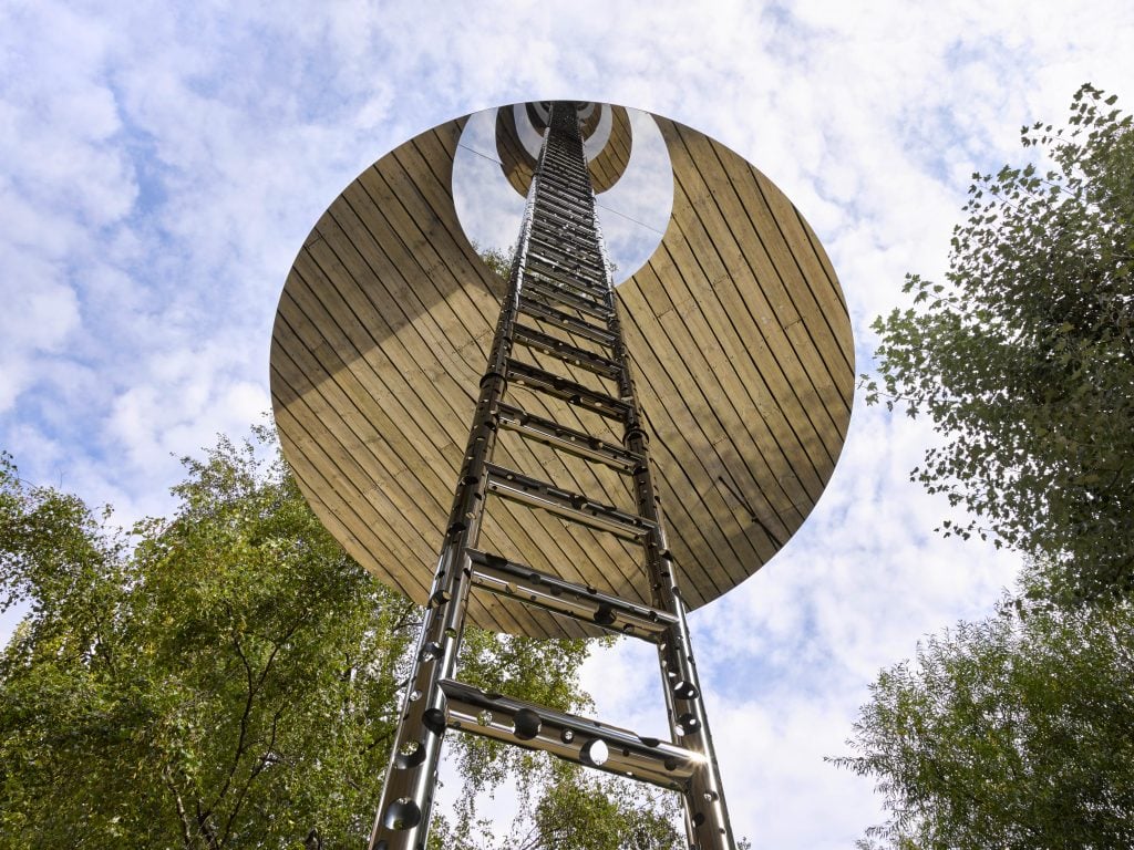 A ladder and the endless reflection of it from the mirrored circular panel, under a sunny sky, with trees