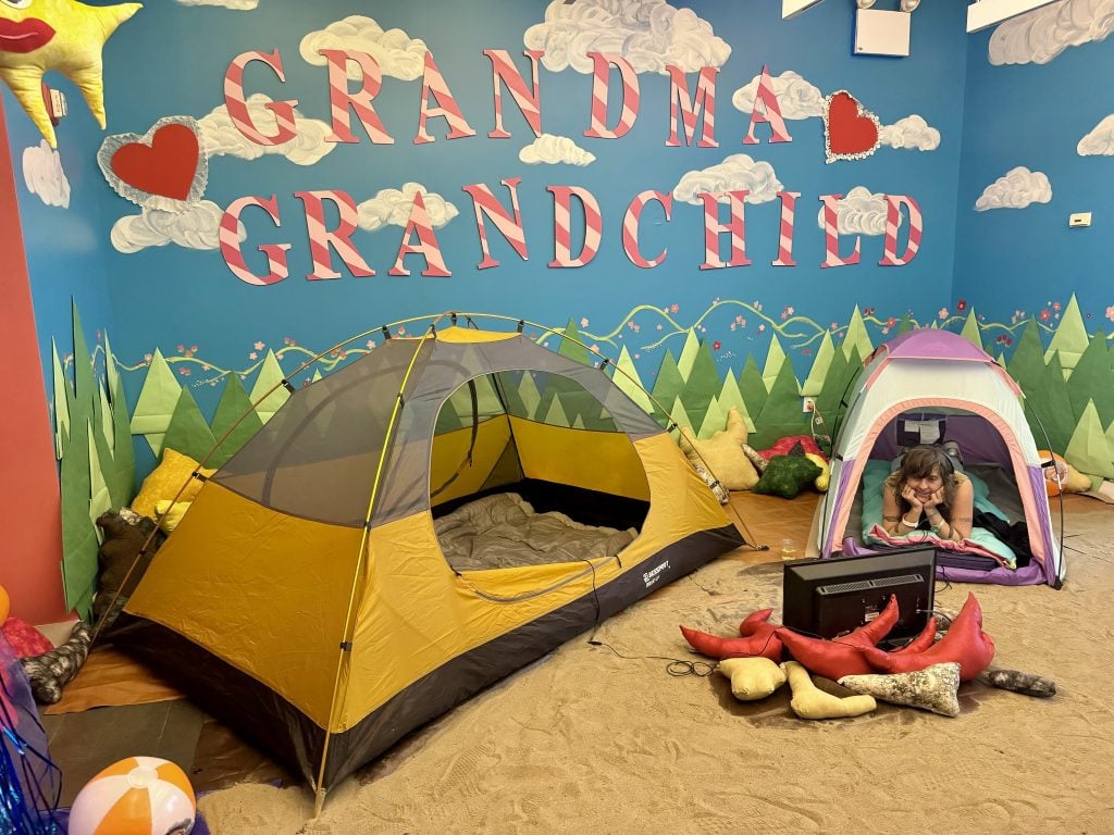 Yoshi Sakai, "Grandma Is Cool AF" curated by Lauren Wolchik at Spring Break Art Show New York 2024. The show title is written on the wall, which is decorated to look like a blue sky with clouds, in red and white striped letters. There are two camping tents on the floor of the gallery, with a woman lying down in one facing a television screen set inside a fake campfire. 