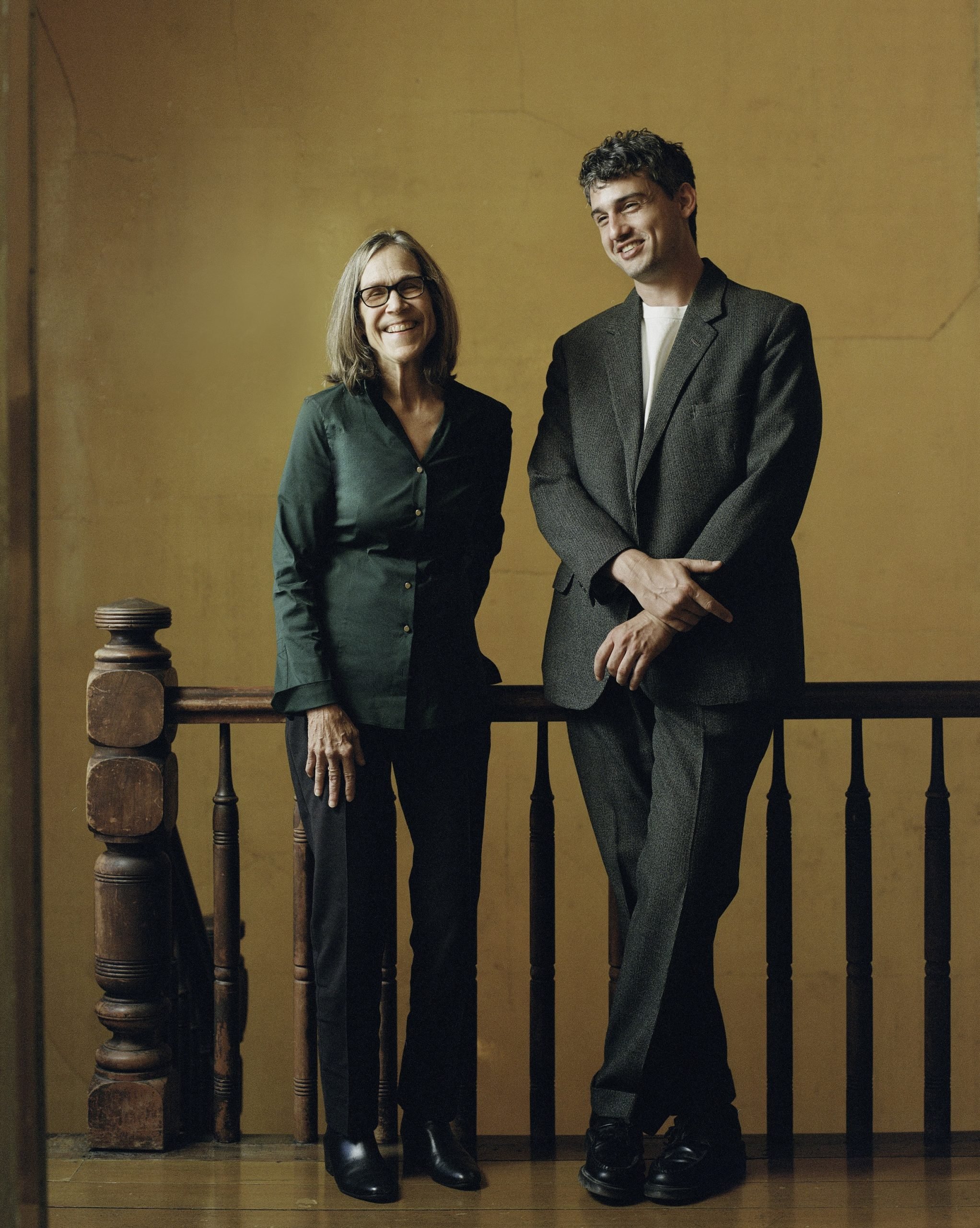 Zully Adler and Robin Wright, director and founder of the Further Triennial in Northern California. The two are standing against a wooden railing at the top of a staircase, against a mustard-colored wall. She is the older woman on the left, and he is the taller, younger man. Both are wearing pant suits.