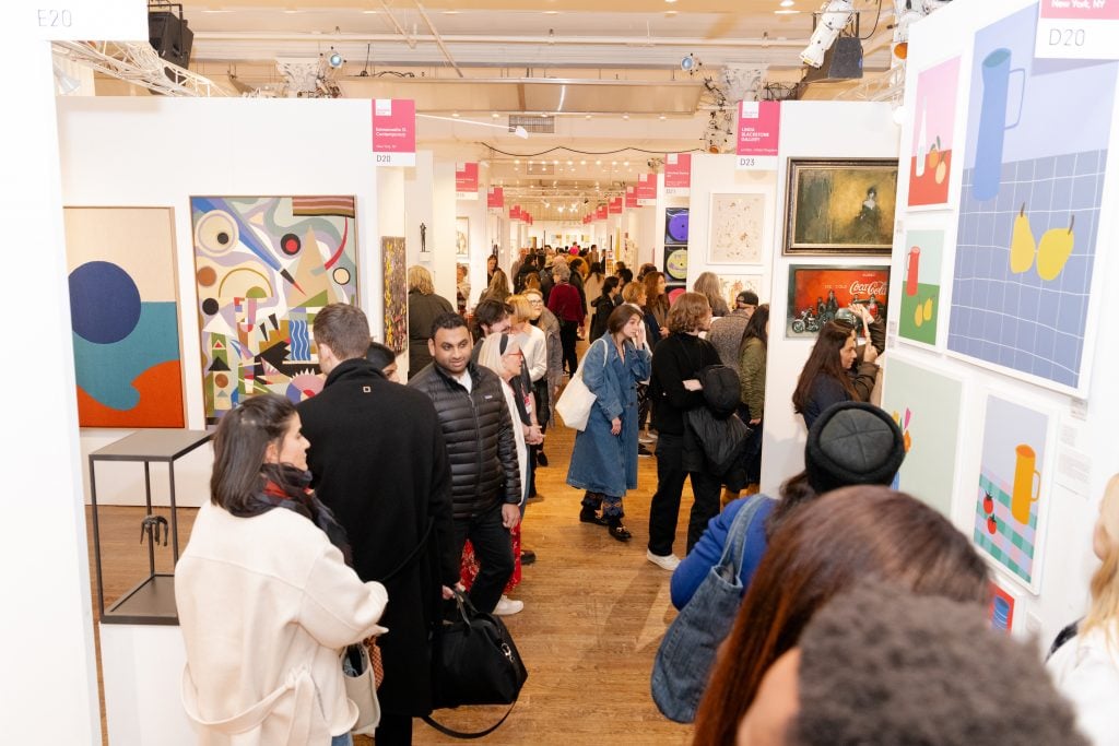 Inside Affordable Art Fair NYC looking down an aisle lined with booths presenting artworks and crowded with visitors.