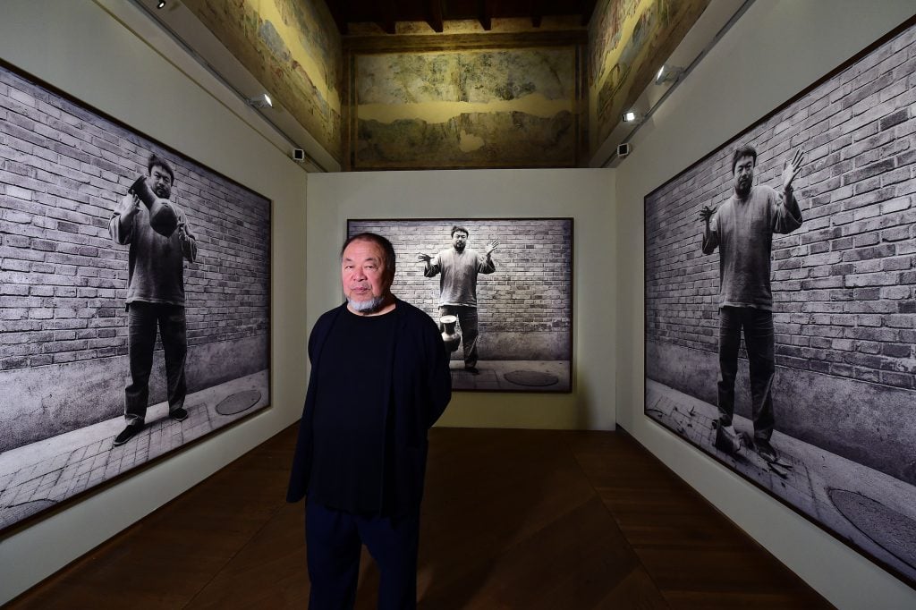 a man wearing an all-black outfit stands in a gallery space that is lined with large black-and-white photographers of himself as a younger man holding and then dropping a ceramic vase