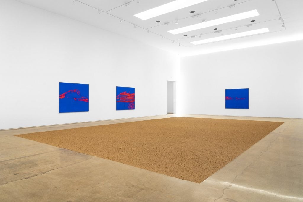 a room with a square installation of granite on the floor and blue paintings with red marks hanging on the wall around it