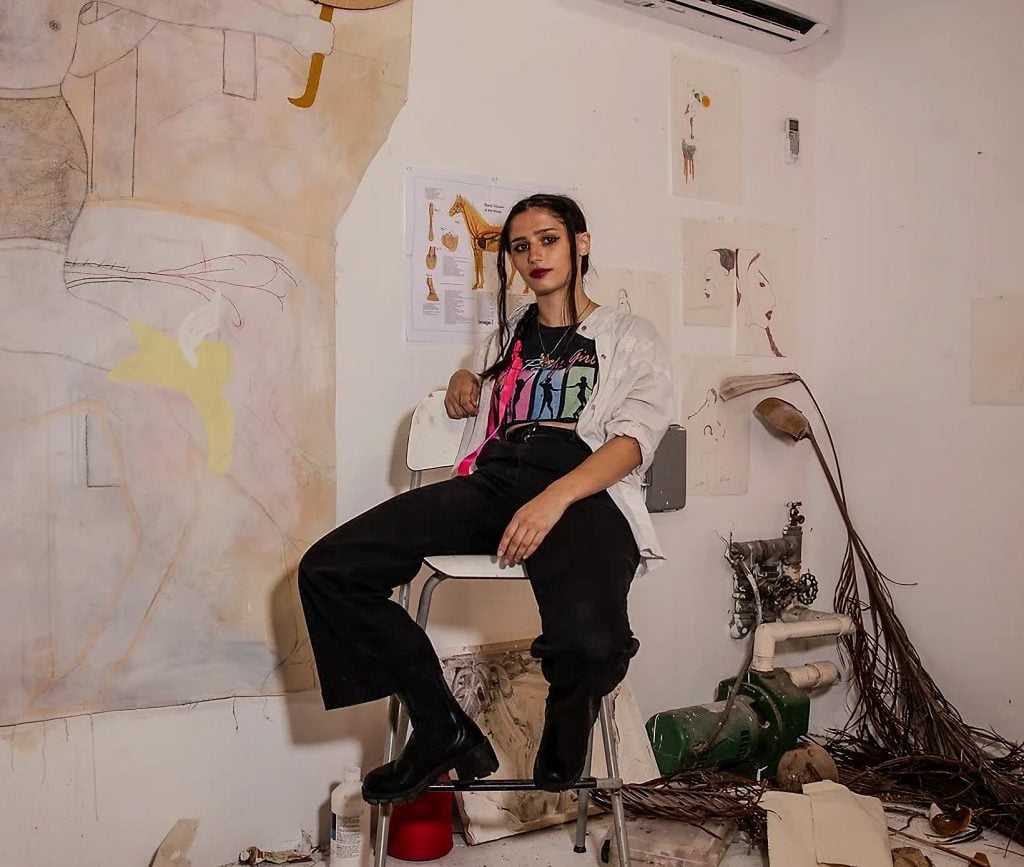 Artist Amy Bravo sitting on a stool that's on a desk in her studio surrounded by in-progress works and found objects.