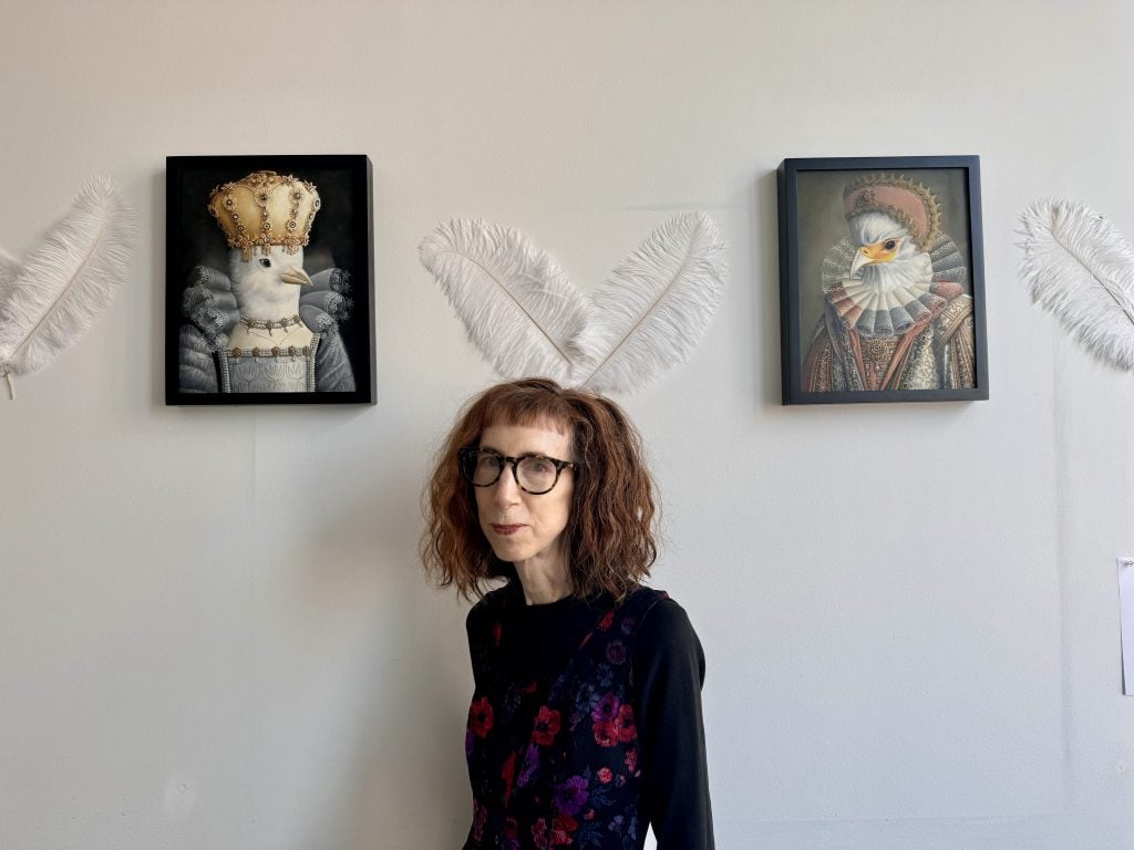 Amy Hill with her "Hybirds" paintings at Spring Break Art Show New York 2024. An older women with shoulder length red wavy hair and glasses stands beneath two white feathers mounted on the wall between a pair of paintings of pigeons dressed in ornate Renaissance clothing. 