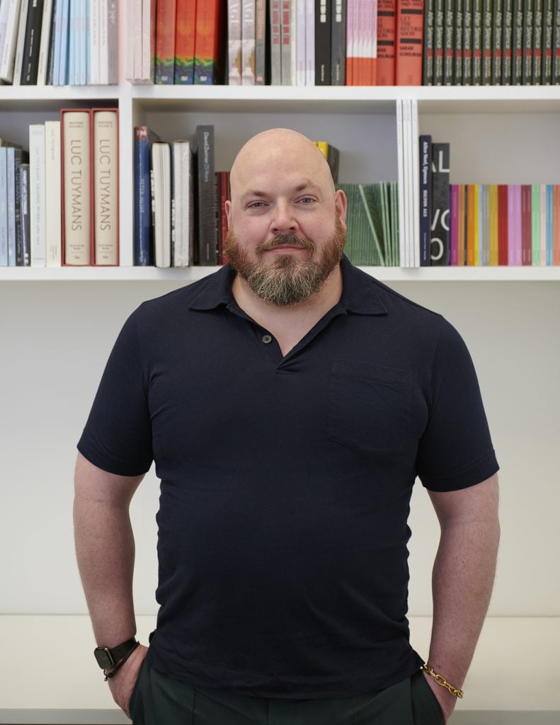 Half portrait of James Morrill, founder of Art Enterprise Resources, wearing a black polo tee standing in front of a bookshelf.