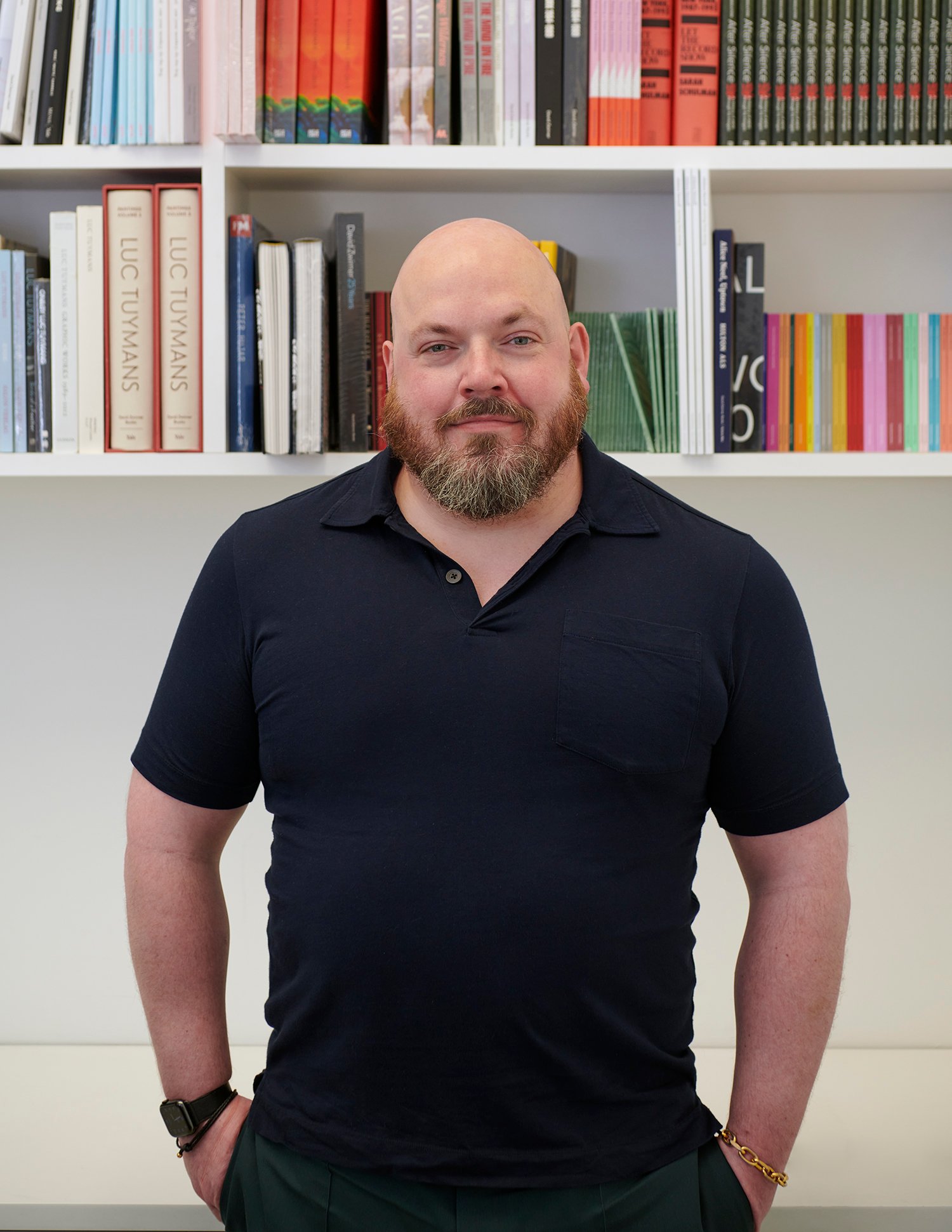 Half portrait of James Morrill, founder of Art Enterprise Resources, wearing a black polo tee standing in front of a bookshelf.