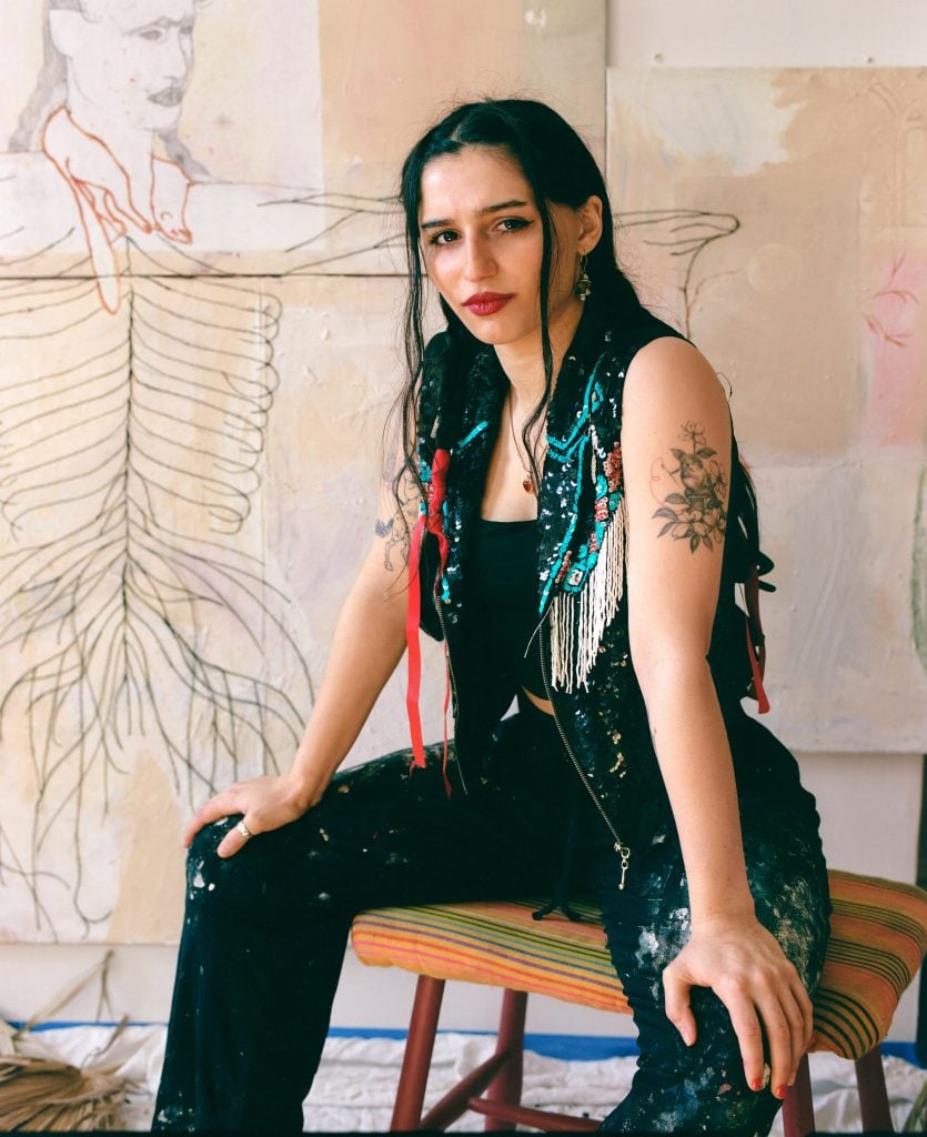 Portrait of artist Amy Bravo wearing mostly black with braided hair sitting on an upholstered square stool in front of some of her paintings in the studio.