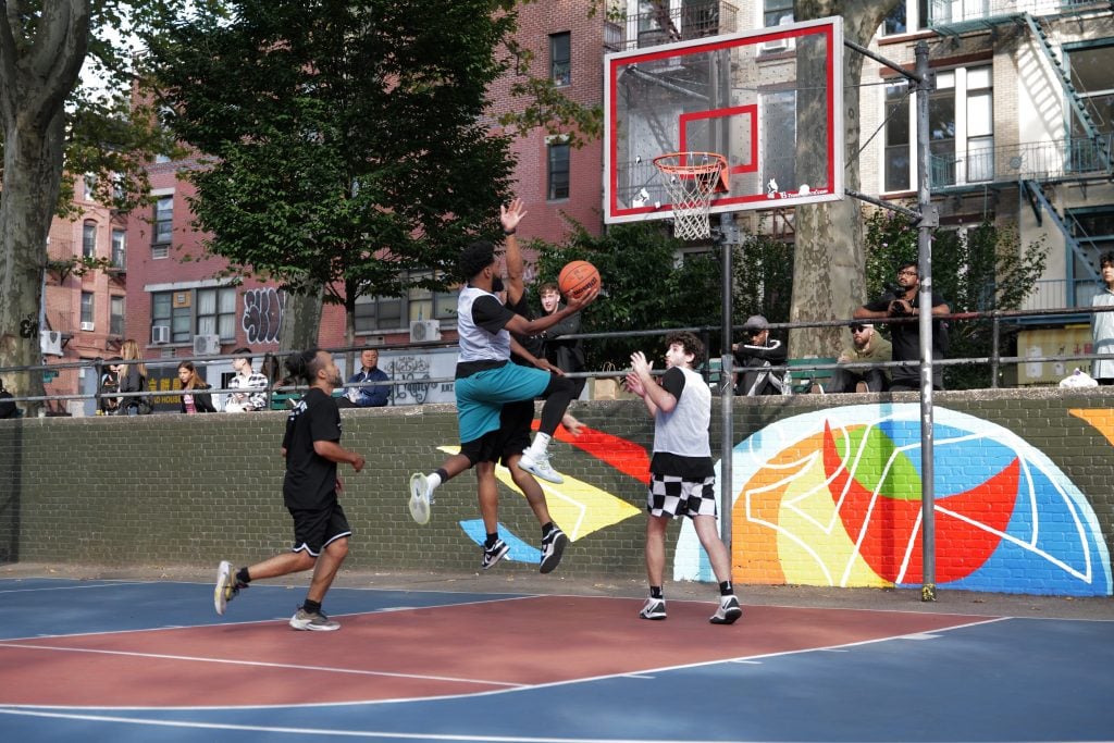 A photograph of a outdoor basketball game in action, with one player flying through the air to make a shot.