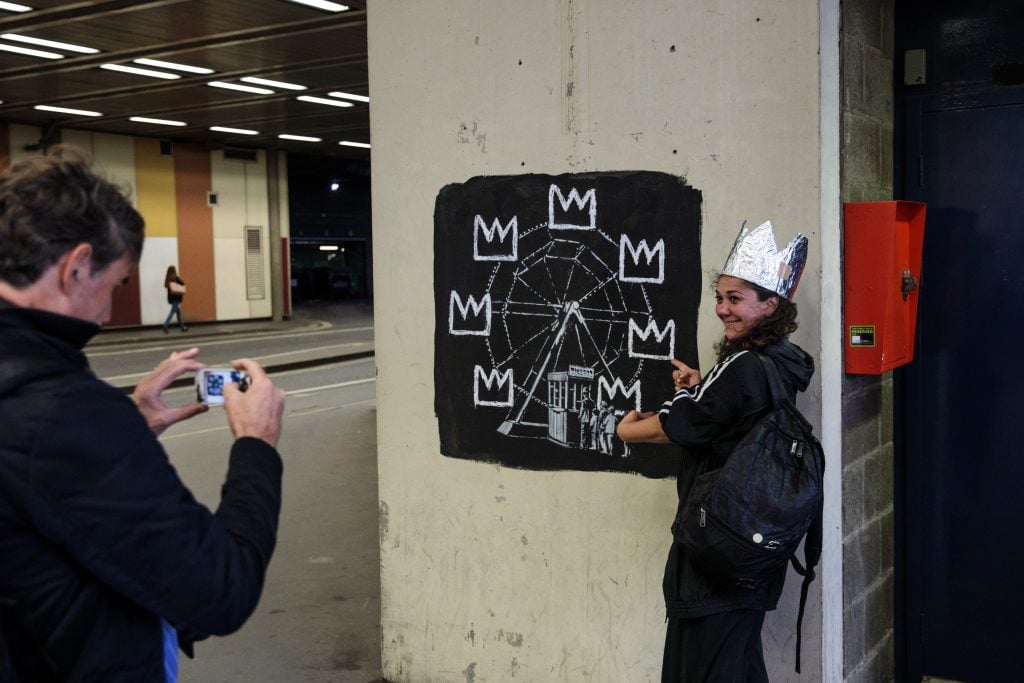 A member of the public takes a photograph a woman wearing a foil crown next to a new work by street artist Banksy on a wall by the Barbican Center in London, England. The two new Banksy murals which have appeared mark the opening of an exhibition by the late American painter Jean-Michel Basquiat at the museum. The pictured mural shows crowds lining up to get tickets for a ferris wheel on a black background. Basquiat's signature crowns take the place of the carts.