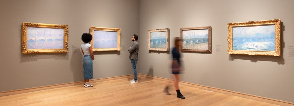 people mill around around a gallery space with hazy landscape paintings hanging on the wall