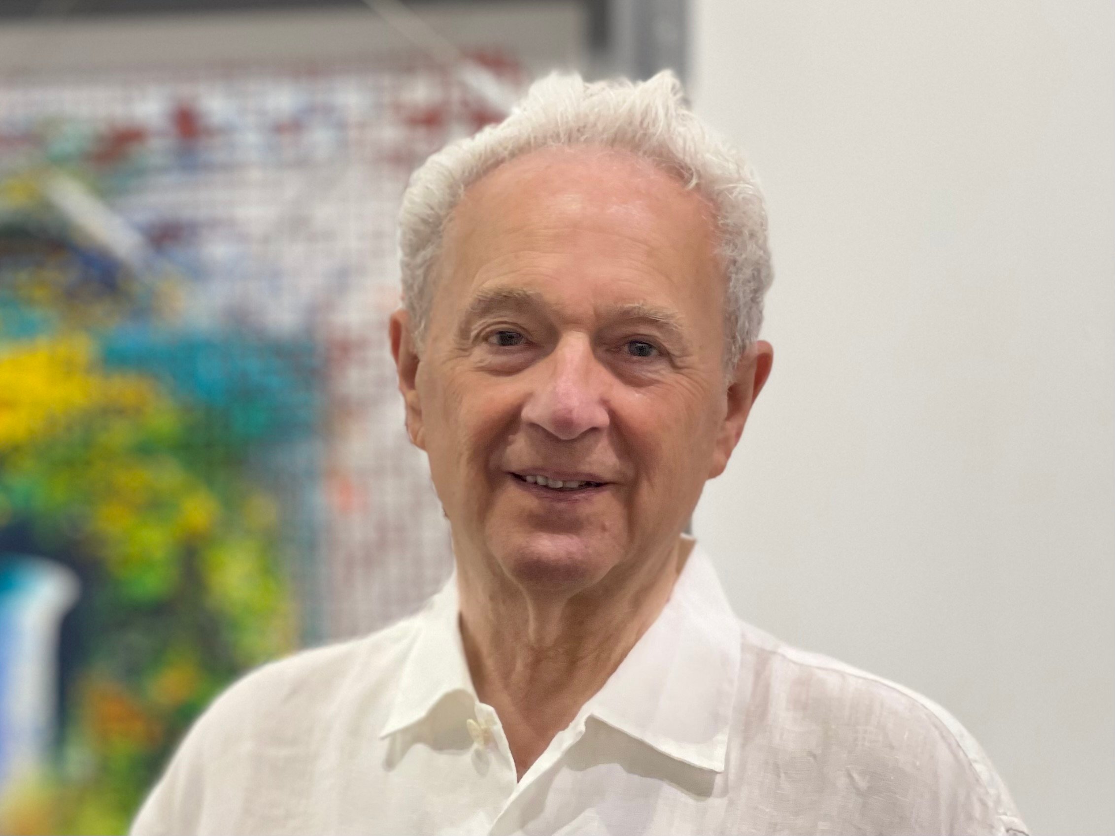 Portrait of Bernard Jacobson Gallery, an older man wearing a white button down shirt with art on the wall behind him.