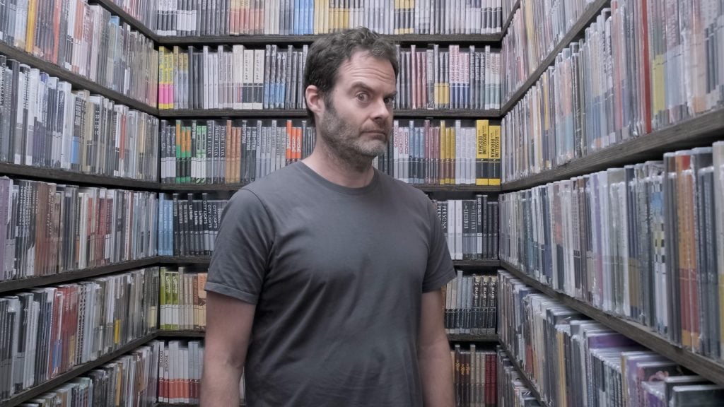 A photo of actor Bill Haderin a gray t-shirt standing with one eyebrow raised. amongst the many films lining the shelves of The Criterion Closet.