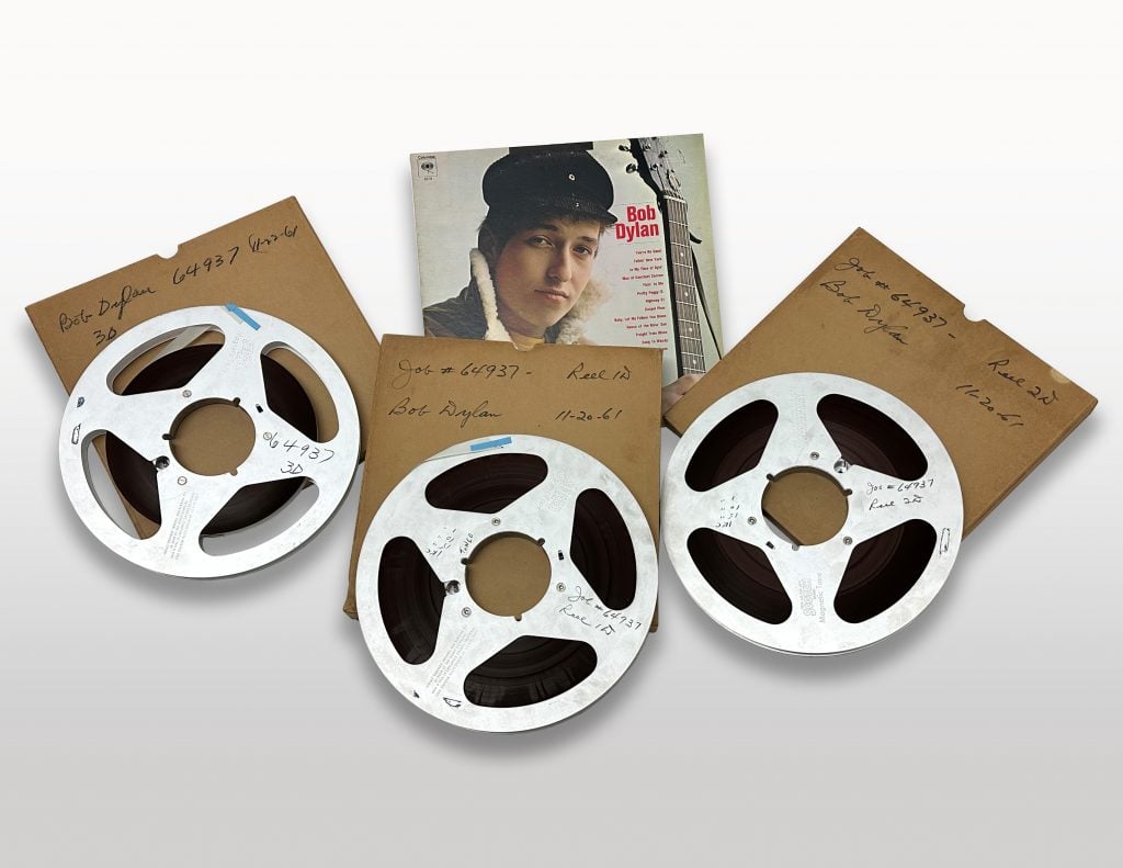 A photo of three old-school tapes atop their original brown slips, with the cover of Bob Dylan's first album in the middle of them, all on a white background.